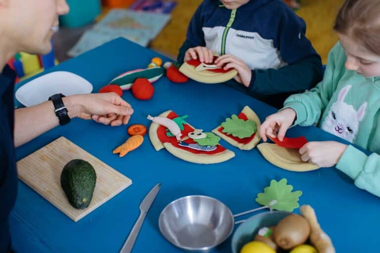 Kinder belegen gemeinsam an einem blauen Tisch eine Spielzeug-Pizza mit buntem Filzgemüse