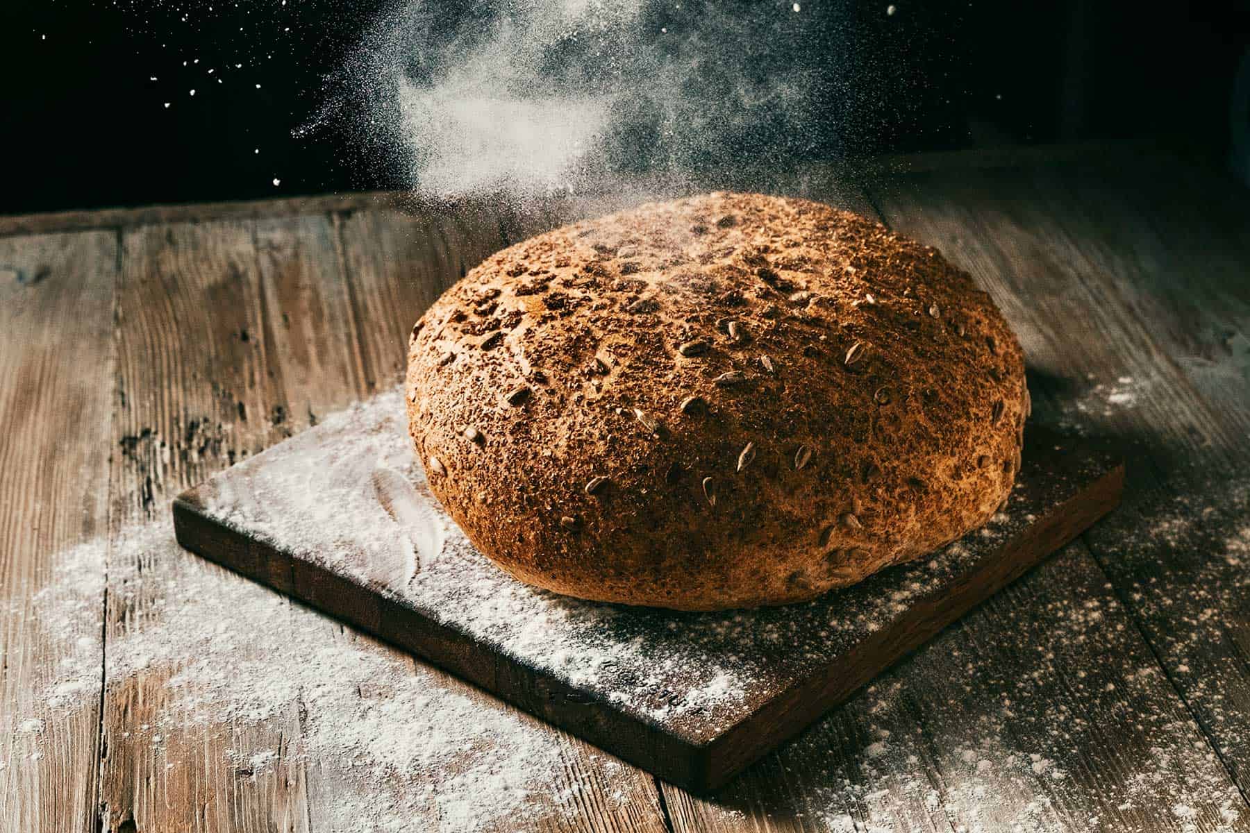 Ein rundes Körnerbrot mit Mehl liegt auf einem Holzbrett