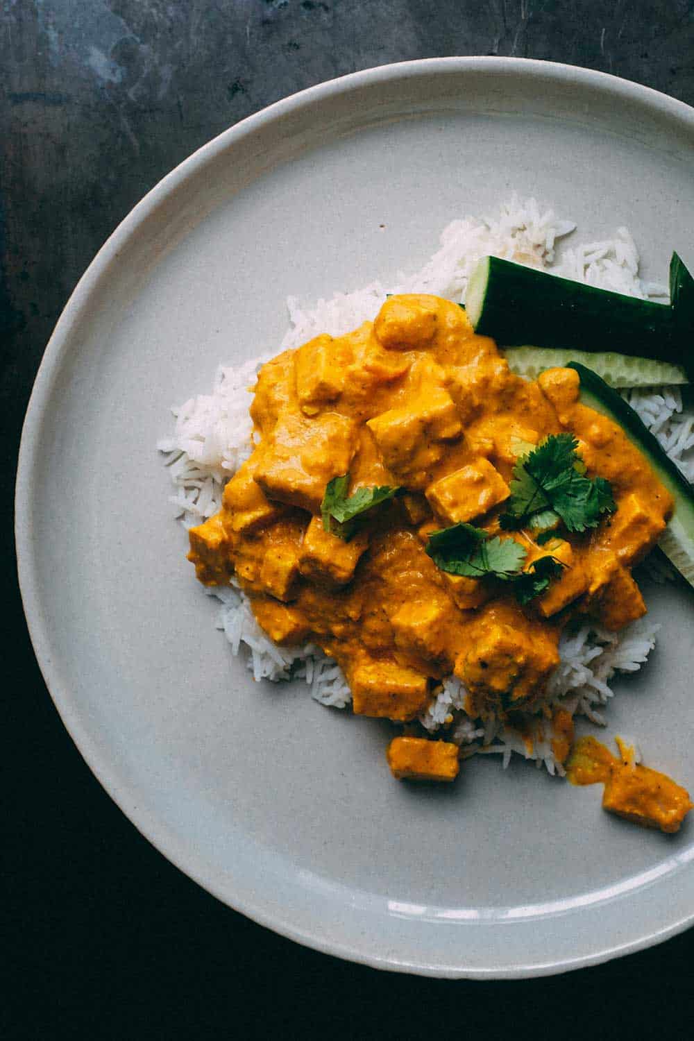 Teller mit Tofu-Curry in gelber Sauce auf weißem Reis, garniert mit Koriander und Gurke